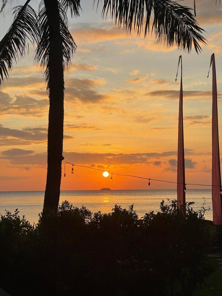Sunrise over the sea framed by palm trees and Balinese penjor poles in the hotel garden
