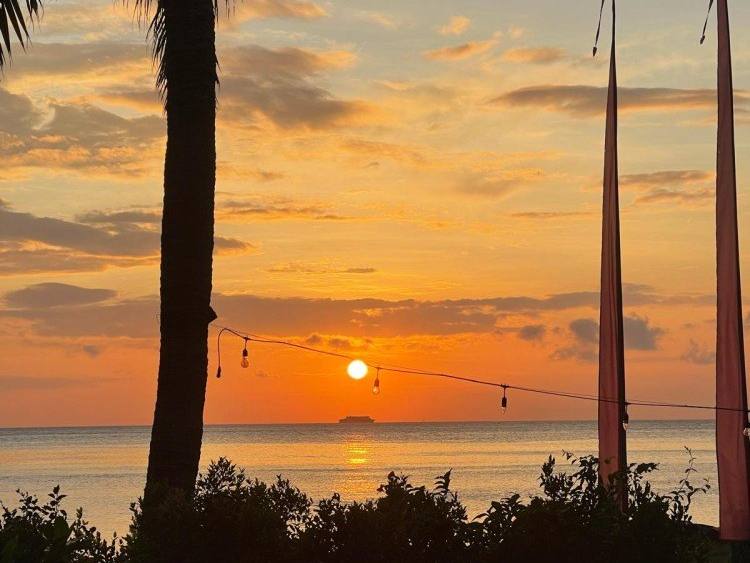 Sunrise over the sea framed by palm trees and Balinese penjor poles in the hotel garden