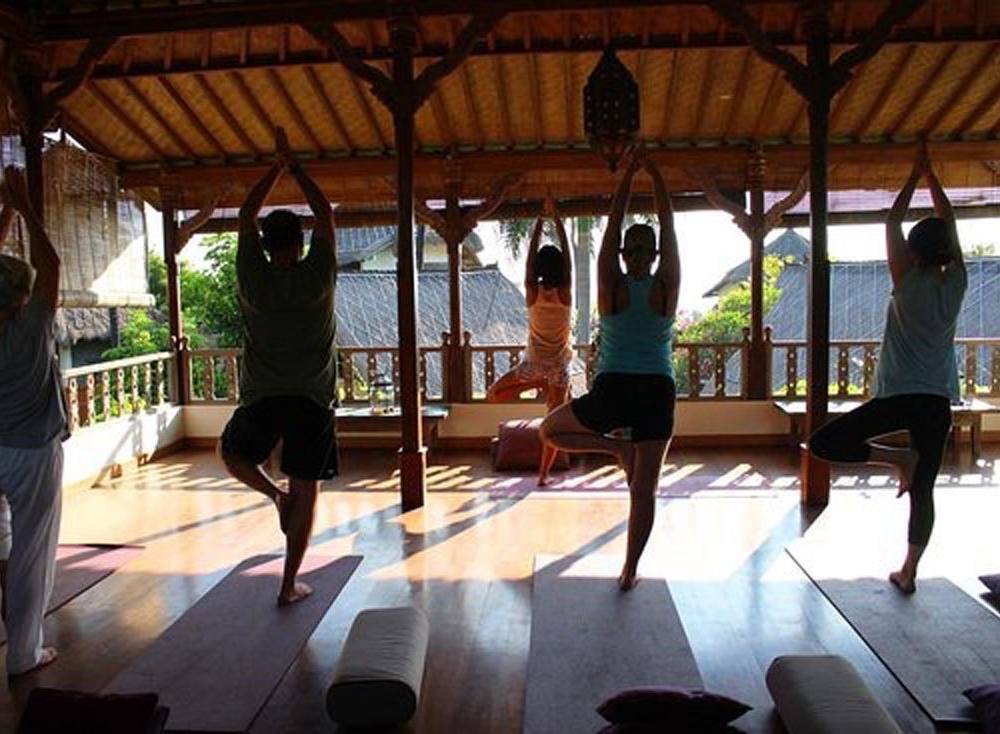 A yoga class in tree pose in the open-sided shala